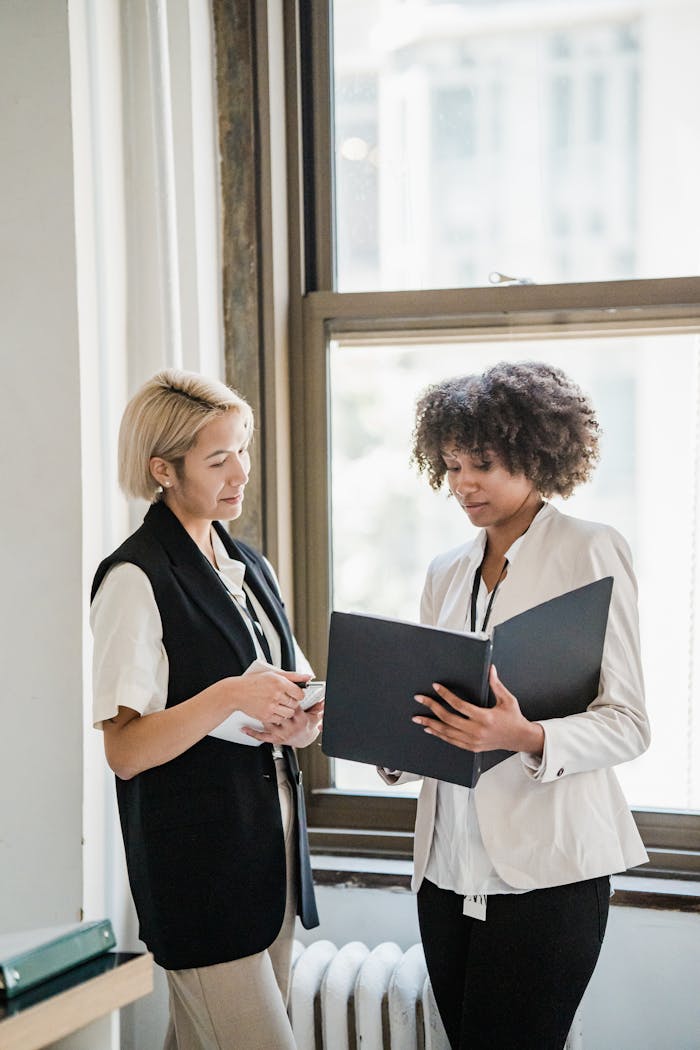 Two women in business attire discussing documents by a window in the office.