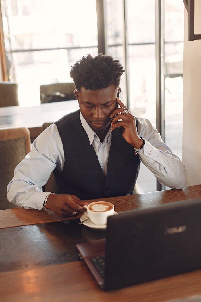 Top view of focused male entrepreneur in formal wear speaking on phone while drinking coffee in front of netbook in creative cafeteria